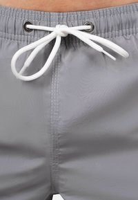 Shorts de bain gris avec une taille élastique et un cordon blanc. Comprend des œillets en métal et des détails de couture contrastants blancs.