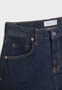 Tamne traperice tamnog denima s klasičnim dizajnom s pet džepova, s narančastim šavovima i sjajnom metalnom kopčom. Vidljiva oznaka veličine.