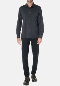 Camicia nera a maniche lunghe con bottoni, realizzata in tessuto liscio, con colletto classico e chiusura frontale a bottoni. Indossata con pantaloni neri a gamba dritta.