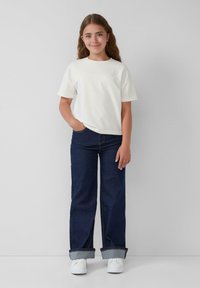 Witte katoenen t-shirt, korte mouwen, losse pasvorm. Donkerblauwe denim jeans met wijde pijpen, omgeslagen bij de enkels. Witte sneakers. Eenvoudig ontwerp.