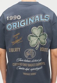 Πίσω μέρος μπλε μπλουζάκι με μεγάλο κείμενο "ORIGINALS", γραφικό τριφύλλι και διάφορες μικρότερες φράσεις όπως "1990", "LIBERTY" και "Certified Daily Basics Garments".