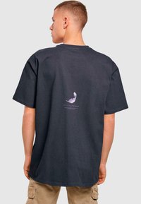 Mørkegrå bomulds T-shirt med en afslappet pasform. Har et lille lilla fjerdesign og tekst trykt på ryggen. Korte ærmer, rund hals.
