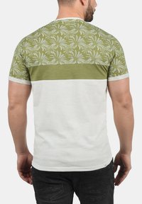 Herret-shirt med et grønt mønstret top med palmebladdesign, en ensfarvet olivenfarvet bånd, og en hvid nederste del. Korte ærmer og rund k neckline.