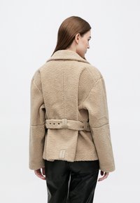 Vrouw draagt een beige, gestructureerde fleecejas met een riem aan de achterkant en zwarte broek, gezien van achteren tegen een effen witte achtergrond.