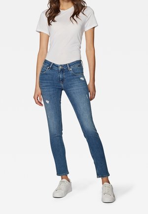 Femme portant un t-shirt blanc à manches courtes, un jean slim bleu légèrement usé, et des baskets blanches, debout devant un fond uni.