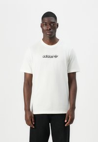 Light beige cotton t-shirt with short sleeves, crew neckline, and a black printed Adidas logo across the chest. Simple and unadorned design.