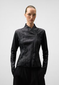 Blusa nera strutturata con maniche lunghe, caratterizzata da pieghe verticali, colletto alto e una texture di tessuto liscia e leggermente lucida.