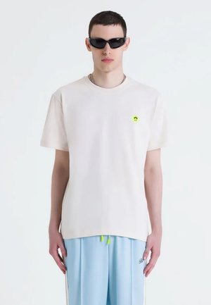 T-shirt beige chiaro a girocollo in cotone con maniche corte, dotata di un piccolo dettaglio grafico verde sul petto. Abbinata a pantaloni blu.