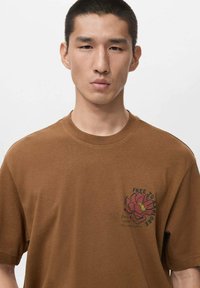 Brązowy bawełniany t-shirt z grafiką różowego kwiatu i napisem „FREE TO EXPLORE” na lewej piersi. Klasyczny fason z okrągłym dekoltem i krótkimi rękawami.