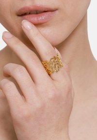 Bague en métal doré avec un design en dentelle complexe ressemblant à des branches. Portée sur un doigt, mettant en valeur sa texture délicate et sa forme.