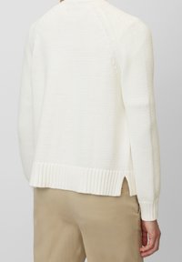 Persona che indossa un maglione bianco a maglia con orlo a coste e spacco laterale, abbinato a pantaloni beige, mostrato di spalle.