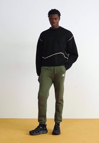 Sort sweatshirt med hvidt bølget linjemønster, parret med grønne sweatpants og sorte sportsko. Enkel, moderne design.