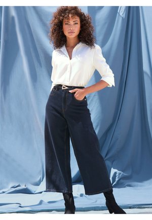 Wide-leg dark denim pants with visible stitching, paired with a white button-up shirt. The model wears a black belt with a triangular buckle.
