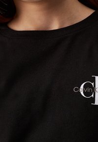 Černé bavlněné tričko s kulatým výstřihem. Na levé straně je bílé logo s nápisem "Calvin" vytištěné moderním písmem. Jemná, hladká textura.