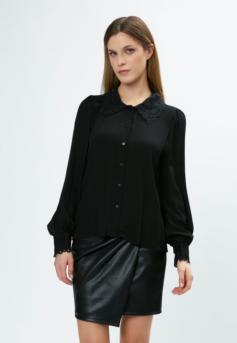 Blusa nera con colletto in pizzo, maniche lunghe a palloncino e chiusura a bottoni. Abbinata a una gonna in pelle nera con design avvolgente.