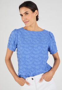 Blauwe geborduurde blouse met korte mouwen, voorzien van een gestructureerd bloemenpatroon en een ronde hals. Gecombineerd met witte broek.