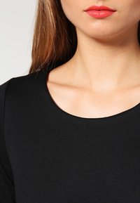 Haut noir à manches longues avec un col rond, texture lisse et coupe ajustée, accentué par des lèvres et des cheveux naturels.