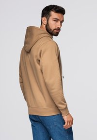 Felpa marrone con cappuccio regolabile e polsini a costine, abbinata a jeans blu. Il tessuto sembra morbido e testurizzato. Il design è casual e aderente.