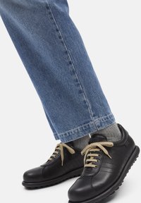 Person wearing light blue jeans, gray socks, and black leather sneakers with beige laces standing on a white background.