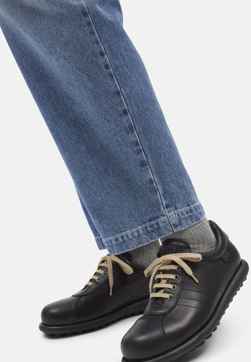 Person wearing light blue jeans, gray socks, and black leather sneakers with beige laces standing on a white background.