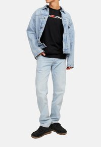 Hellblaues Jeansjacke über einem schwarzen T-Shirt mit weißem Logo, kombiniert mit hellblauen Jeans und schwarzen Sneakern mit strukturierter Oberfläche.