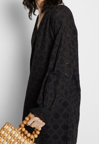 Donna che indossa un abito in pizzo nero con maniche a palloncino, mentre tiene una borsa di legno con perline per il manico rotondo.
