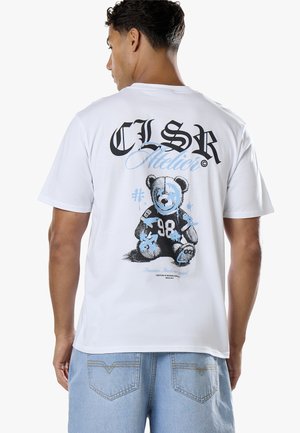 Homme portant un t-shirt blanc avec un graphisme noir et bleu représentant un ours en peluche et le texte "CLSR Atelier" dans le dos ainsi qu’un jean bleu clair.