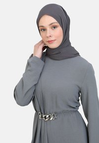 Šedé šaty s dlouhými rukávy a úzkým pasem, zdobené stříbrným řetízkem. Tkanina má hladkou strukturu a jemný drapérii.