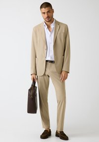 Beige pak met inkeping revers, lange mouwen en twee voorzakken. Gecombineerd met een wit overhemd, bruine riem en donkerbruine leren tas.