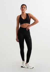 Svart sports-BH og høytlivsleggings laget av elastisk stoff, med et elegant design og en logo på benet. Brukes med hvite joggesko.