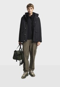 Crna vodonepropusna jakna s kapuljačom, uparena s tamnim duksericom, kaki cargo hlačama i maslinasto zelenom torbom. Sadrži čvrste cipele s bež detaljima.