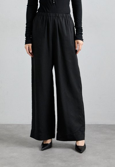Black wide-leg trousers with an elastic waistband, made from smooth fabric. Worn with pointed black heels, featuring a clean, streamlined design.