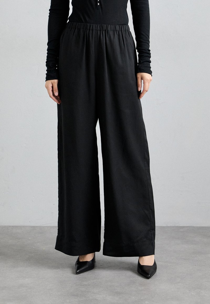 Black wide-leg trousers with an elastic waistband, made from smooth fabric. Worn with pointed black heels, featuring a clean, streamlined design.