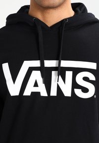 Uomo che indossa un felpa nera con il logo bianco di Vans sul petto, con i lacci della felpa che pendono davanti.