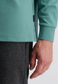 Licht turquoise lange mouwen shirt met een label aan de zijkant, gecombineerd met gestructureerde donkere broek met een subtiel patroon. Close-up van de mouw en de hand.