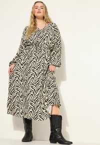 Lang, losvallende jurk met een zebra-print in zwart en beige, aangesnoerd bij de taille, gecombineerd met zwarte cowboylaarzen.