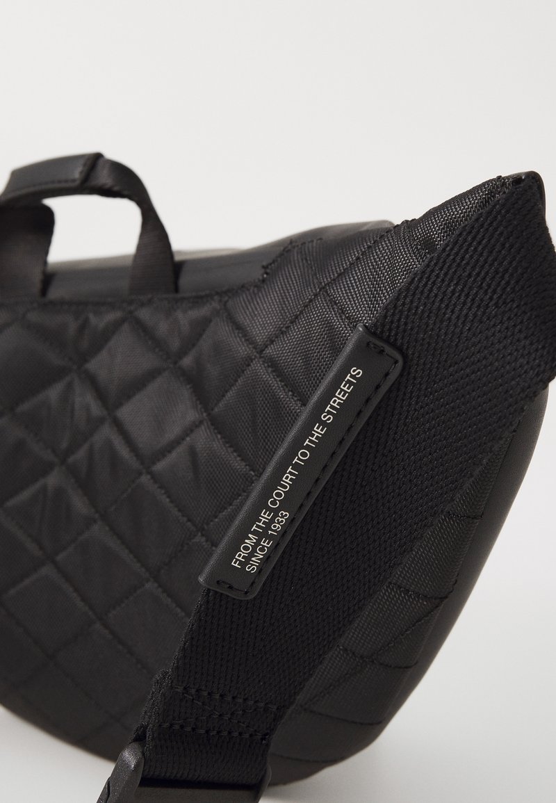 Sac noir matelassé avec une surface texturée, doté d'une poignée supérieure et d'une sangle ajustable. Une étiquette indique : "DU TERRAIN À LA RUE DEPUIS 1933."