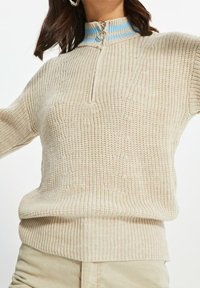Beige gestrickter Pullover mit einem Halb-Reißverschlusskragen und blauen sowie weißen Streifen, strukturiertem Stoff und geripptem Saum.