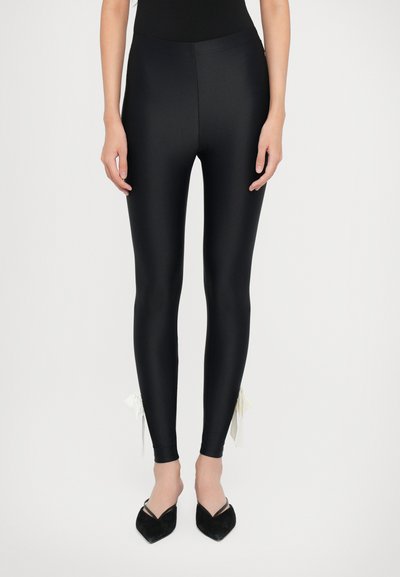 Leggings negros brillantes con diseño ajustado, textura suave y acentos de cinta blanca en el tobillo. Combinados con zapatos de punta negra.