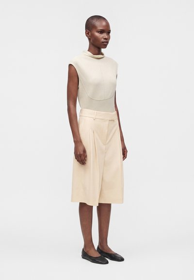 Femme aux cheveux courts portant un haut beige sans manches et un short beige large taille haute avec des chaussures plates noires, debout devant un fond blanc uni.