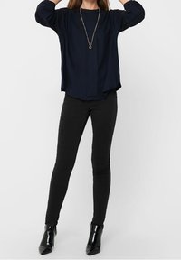 Blusa blu navy a maniche lunghe con dettagli plissettati sulle spalle, abbinata a jeans skinny neri e stivali neri in vernice fino alla caviglia. Accento con una semplice collana dorata.