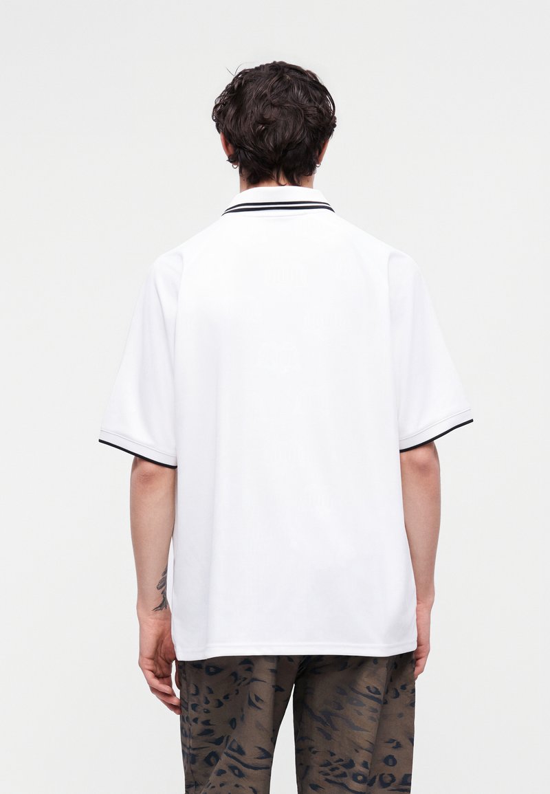Mann mit dem Rücken zur Kamera, trägt ein weißes kurzärmeliges Poloshirt mit schwarzem Besatz und gemusterte braune Hose vor schlichtem Hintergrund.