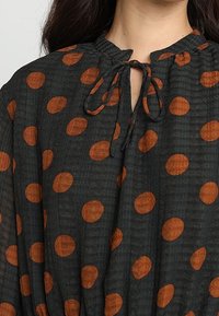 Černá tkaninová blůza s oranžovými puntíky, s uzávěrem na tkaničku u krku a texturovaným povrchem. Navržena pro volnější střih s nařaseným lemem.