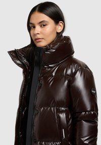 Brown puffer jacket with shiny texture, high collar, snap buttons, and side pocket featuring brand label. Features a fitted silhouette.