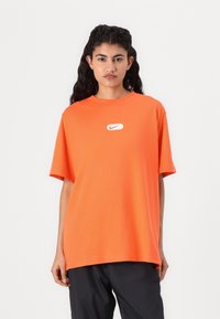 Oranžs kokvilnas T-krekls ar apaļu kakla izgriezumu un īsām piedurknēm. Mazs balts Nike logotips ir centrēts uz krūtīm.