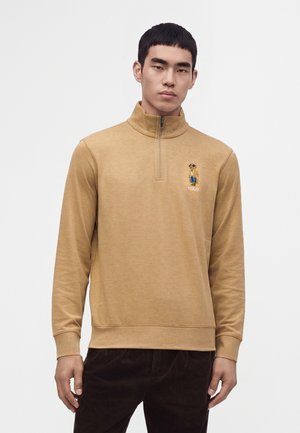 Млад мъж, облечен в светлокафяв суитшърт с quarter-zip, с малко бродирано мече и думата Polo на гърдите.