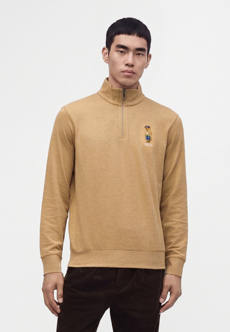 Млад мъж, облечен в светлокафяв суитшърт с quarter-zip, с малко бродирано мече и думата Polo на гърдите.
