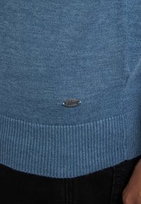 Pulover tricotat de culoare albastru deschis, cu un tiv și mansete cu nervuri, având un mic logo metallic brodat în partea dreaptă jos.