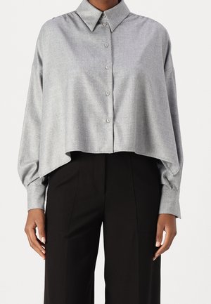 Femme portant une chemise ample gris clair à boutons avec manches longues et un pantalon noir taille haute, debout devant un fond uni.