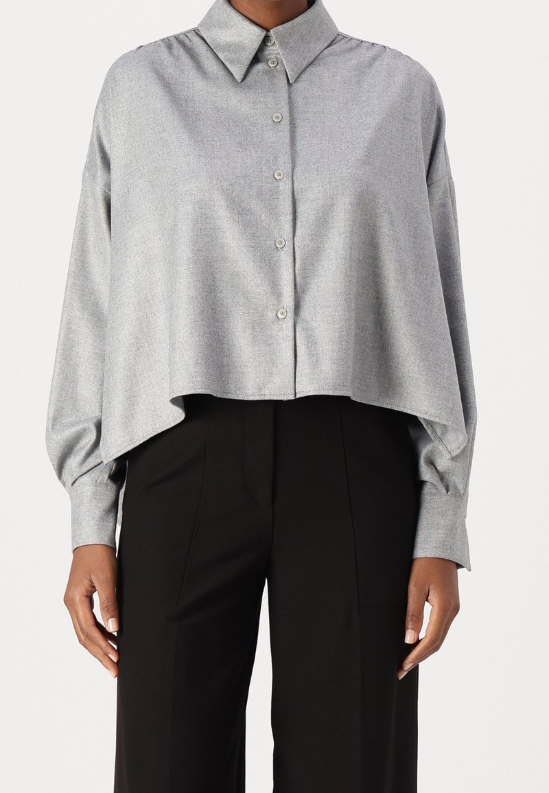 Femme portant une chemise ample gris clair à boutons avec manches longues et un pantalon noir taille haute, debout devant un fond uni.